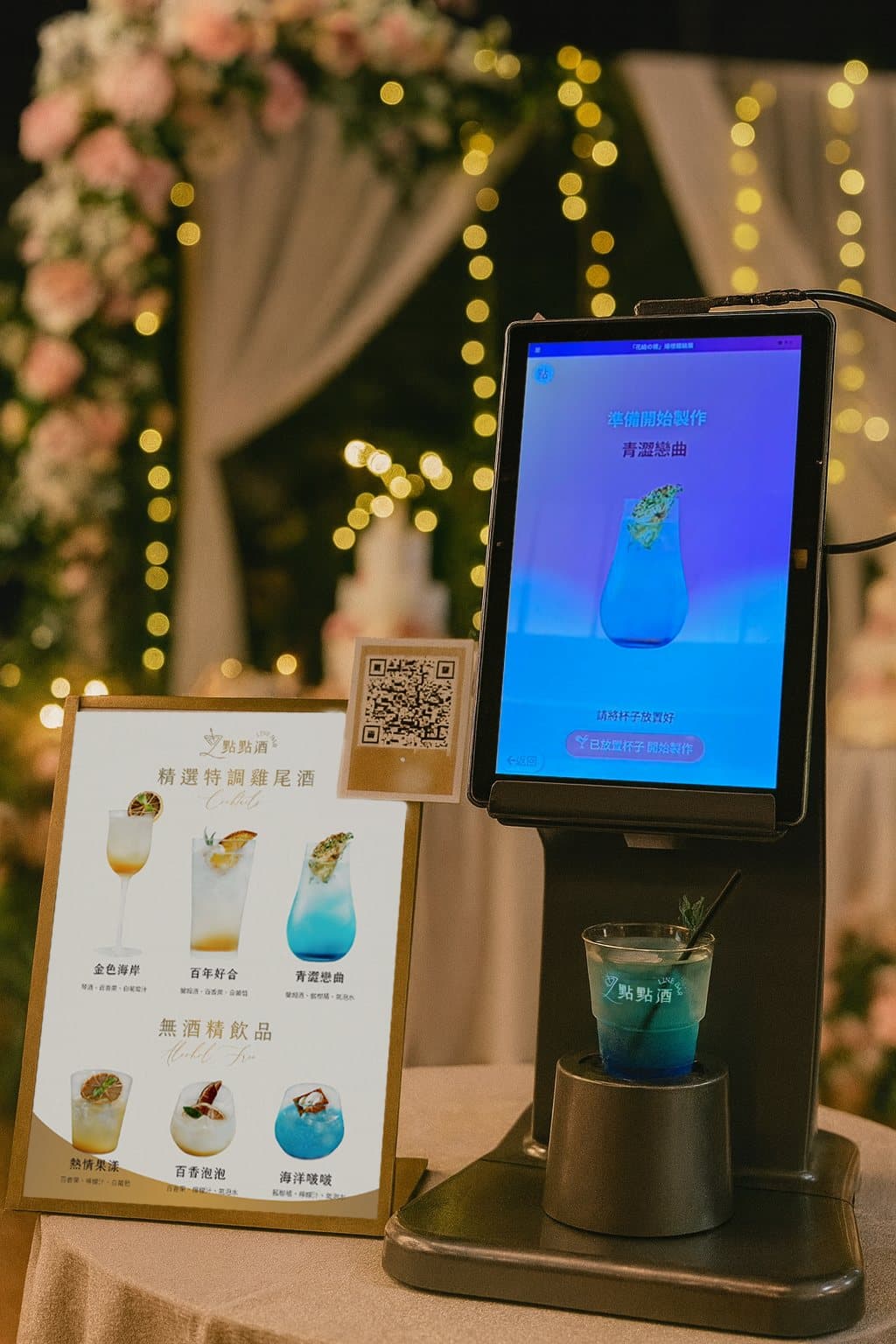 點點酒機台嵌在婚宴的花藝與燈串佈景中,旁邊立著酒單卡與掃碼點酒說明