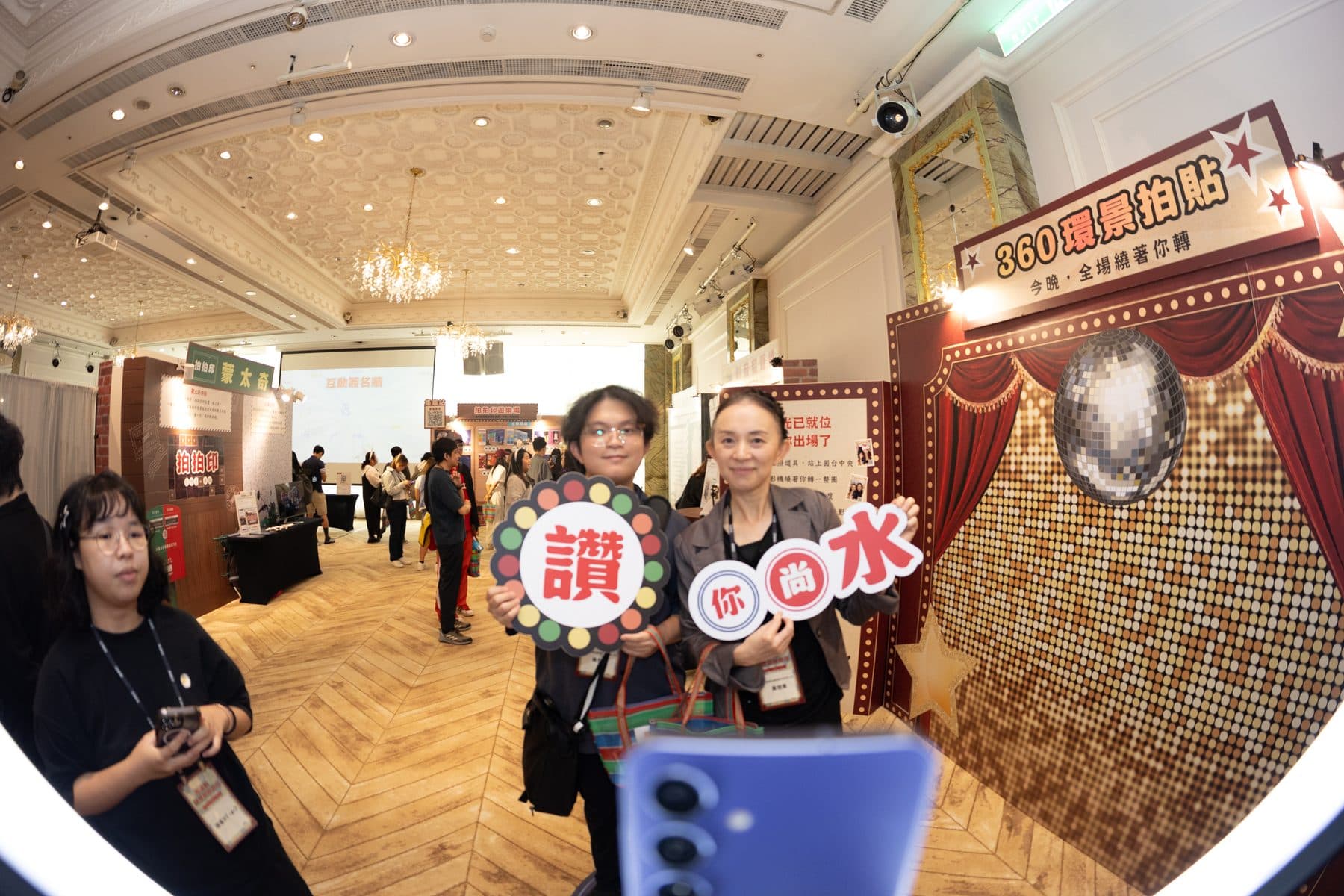 兩位賓客手持「讚」與「你尚水」字樣手舉牌,在 360 環景拍貼背板前合影