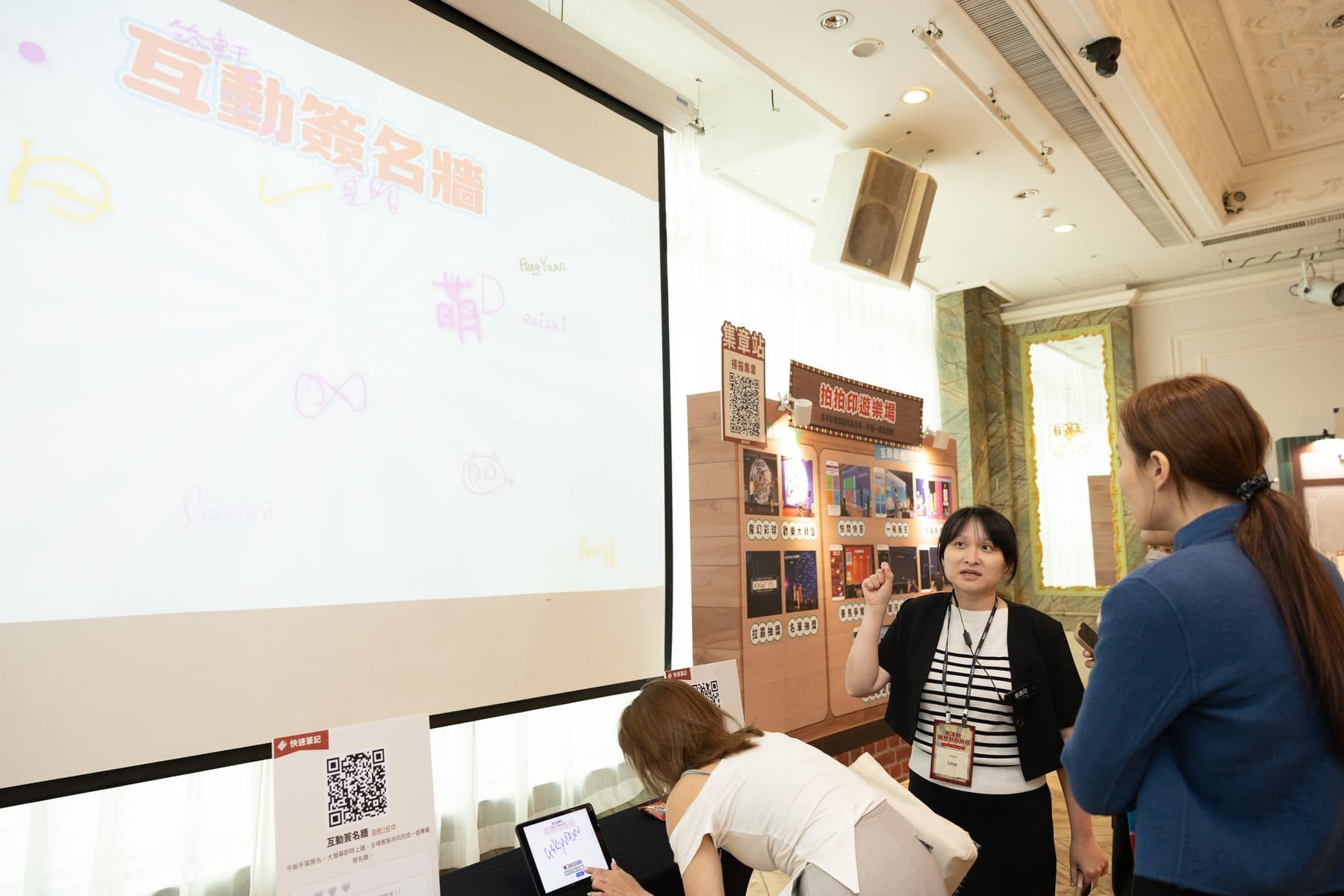 工作人員在投影布幕旁向賓客介紹互動簽名牆,幕上已有多筆手寫簽名