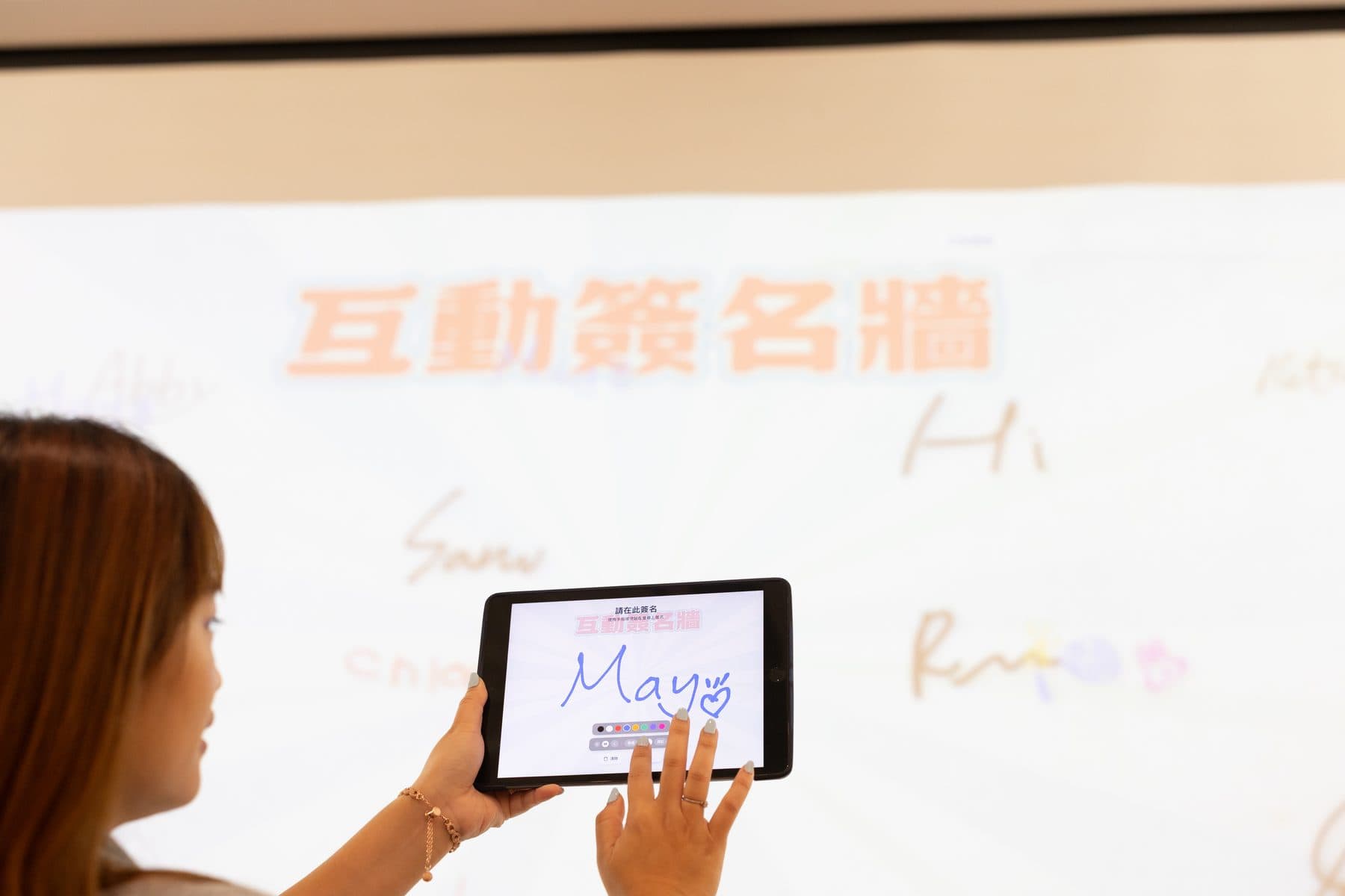 來賓在平板上手寫簽名「May」,後方布幕上已投影出互動簽名牆與其他人的簽名